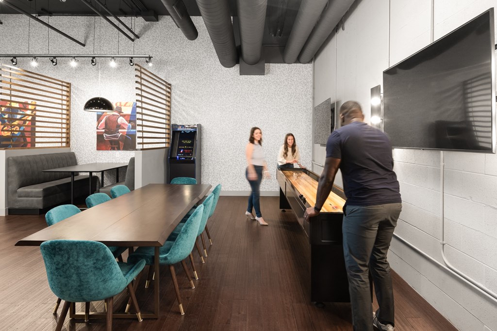 A community lounge with a game center and a table surrounded by eight blue chairs on hardwood-style flooring.