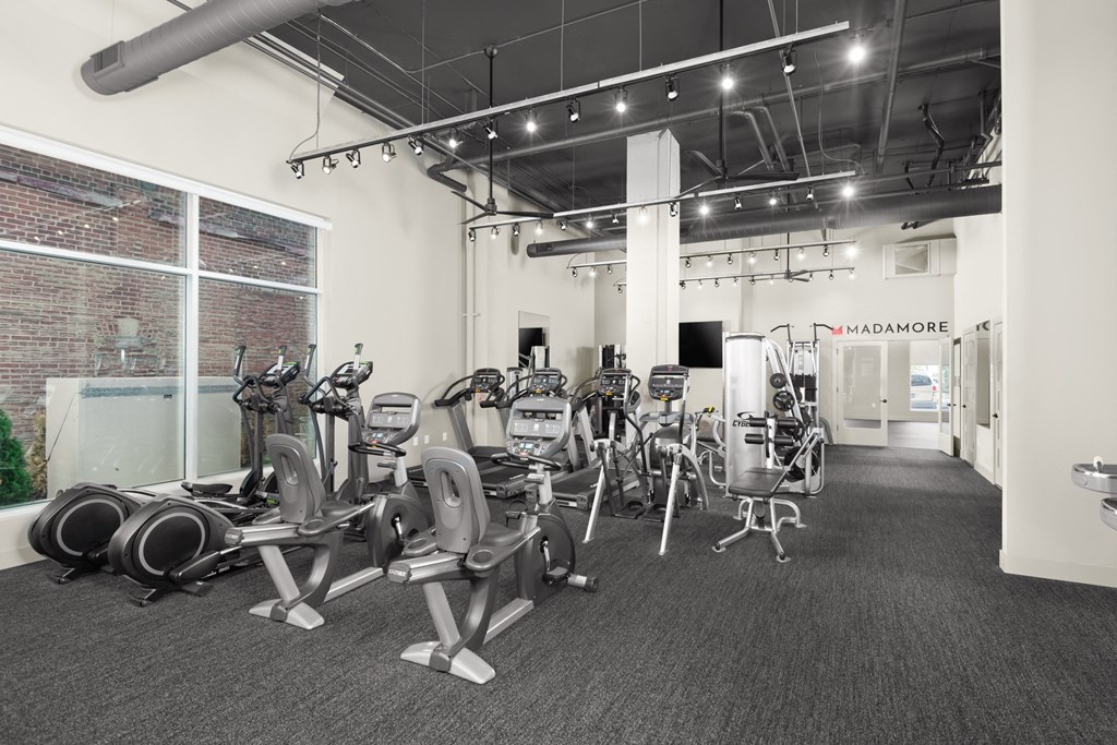 A fitness center equipped with a selection of workout machines on carpeted flooring, and large windows.