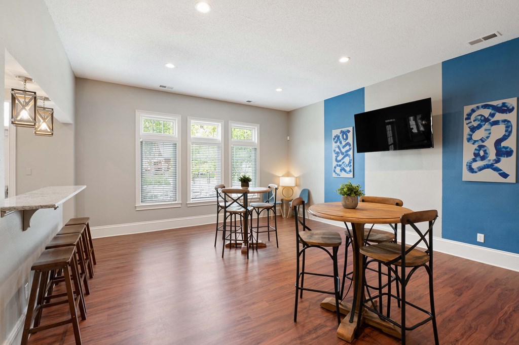 Community clubhouse with two tables each surrounded by three chairs, four barstools, a TV, and windows.