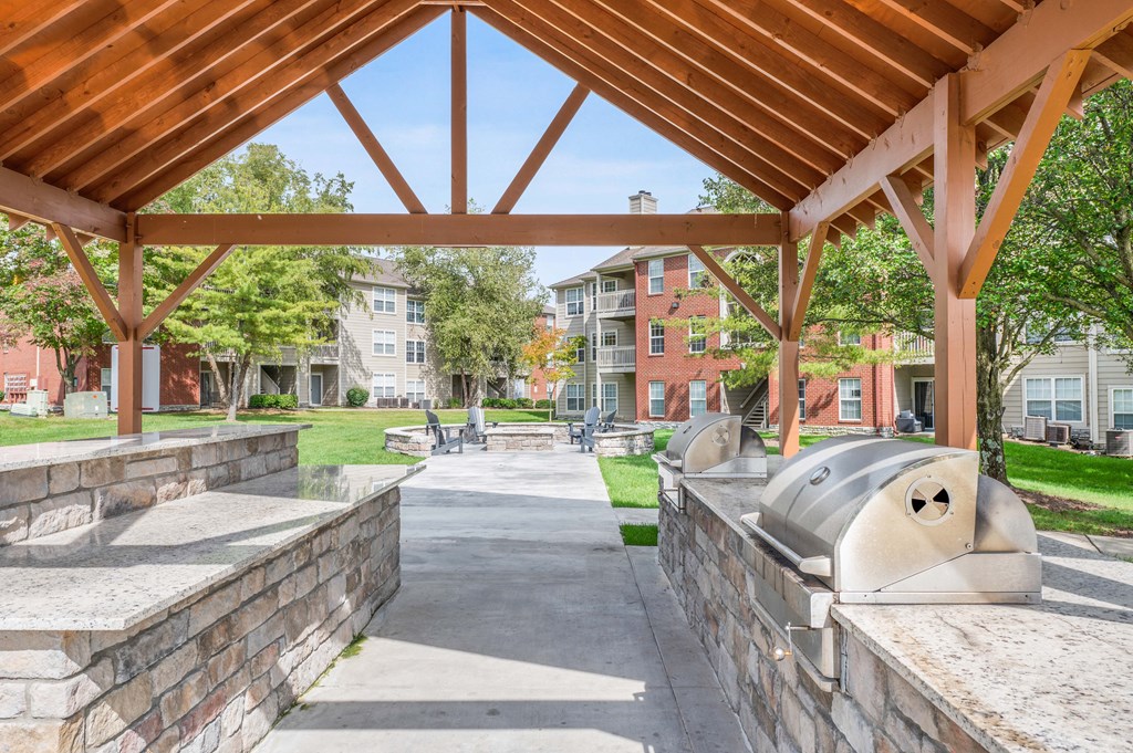 A BBQ area with two grills, next to a firepit surrounded by chairs, next to apartment buildings.