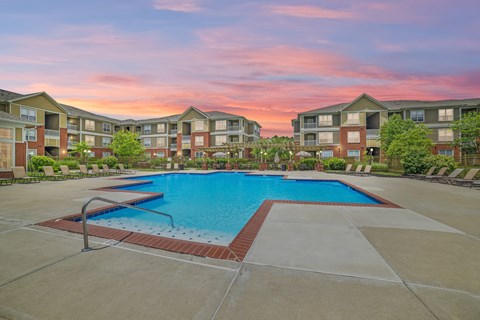 Swimming pool surrounded by chairs, next to apartment buildings, and a sunset.