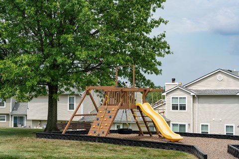 A playground with a yellow slide, a large tree, and grass.