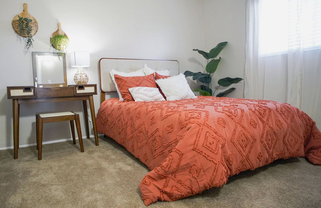 A bed with a red comforter and a white headboard sits in a room with a wooden nightstand and a plant.
