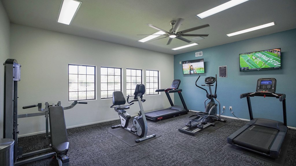 A gym room with a row of treadmills, a fan, and a television showing a baseball game.