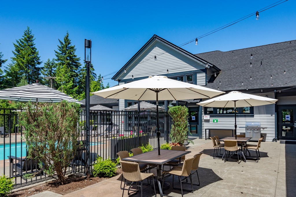 A poolside area with a table and chairs, umbrellas, and a building in the background.