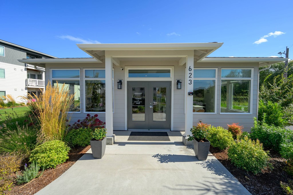 A white building with a glass door and windows is surrounded by plants.