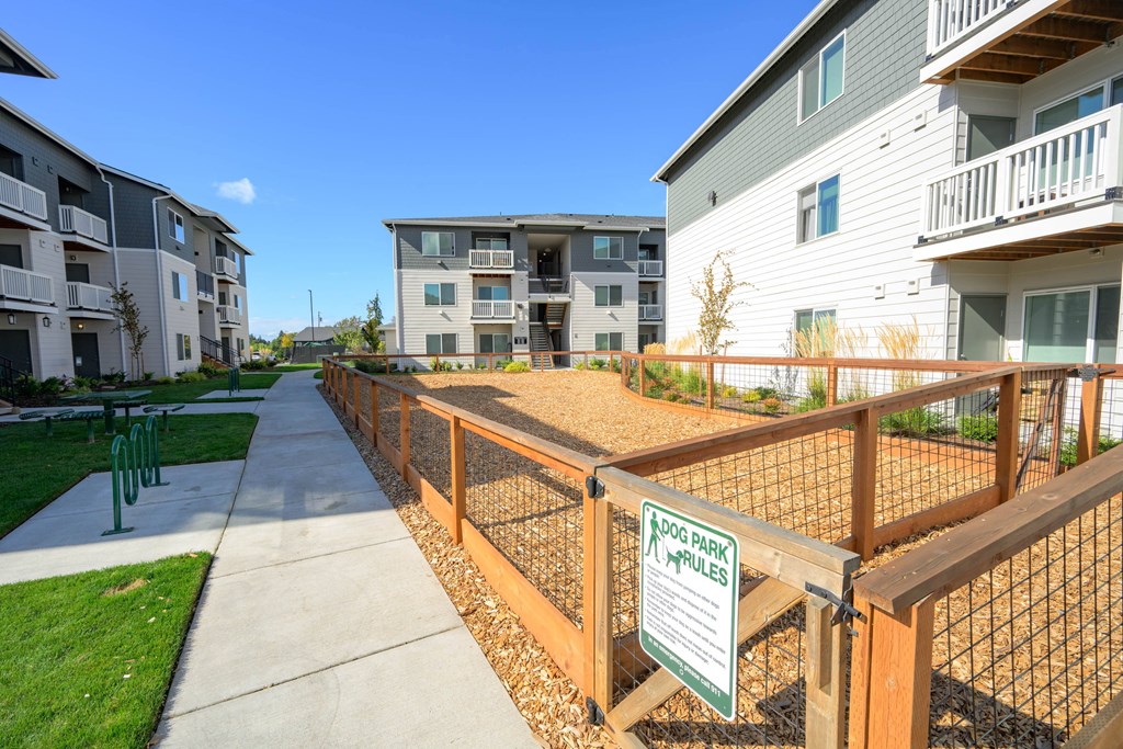 A dog park sign is on a fence in front of apartment buildings.