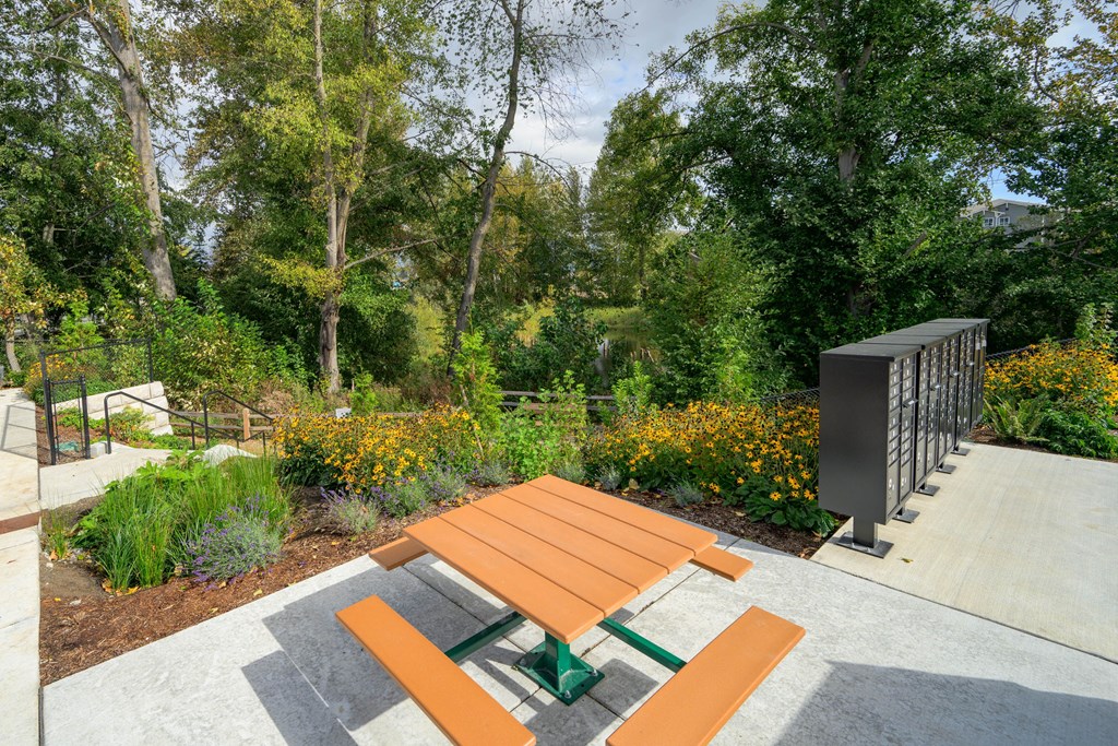A wooden picnic table with a green bench in the middle of a concrete area with a fence and trees in the background.