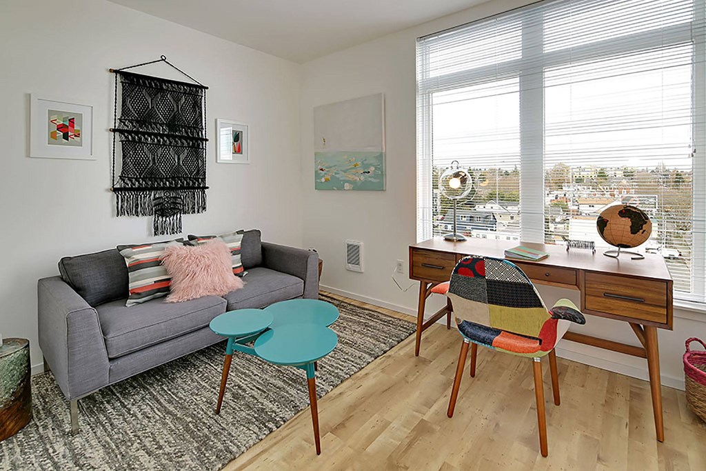 A room with a grey couch, a table, a chair and a window.