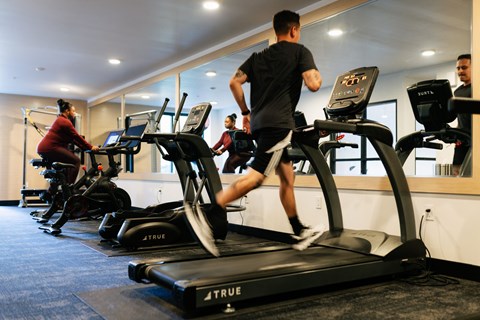 A man is running on a treadmill in a gym with other people using exercise machines.
