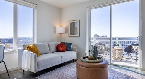 A living room with a grey couch, a small table with a plant on it, and a view of the city through the windows.