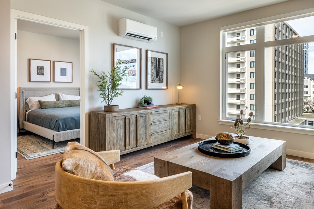 A well-lit, modern bedroom with a large bed, wooden furniture, and a view of a cityscape through a large window.