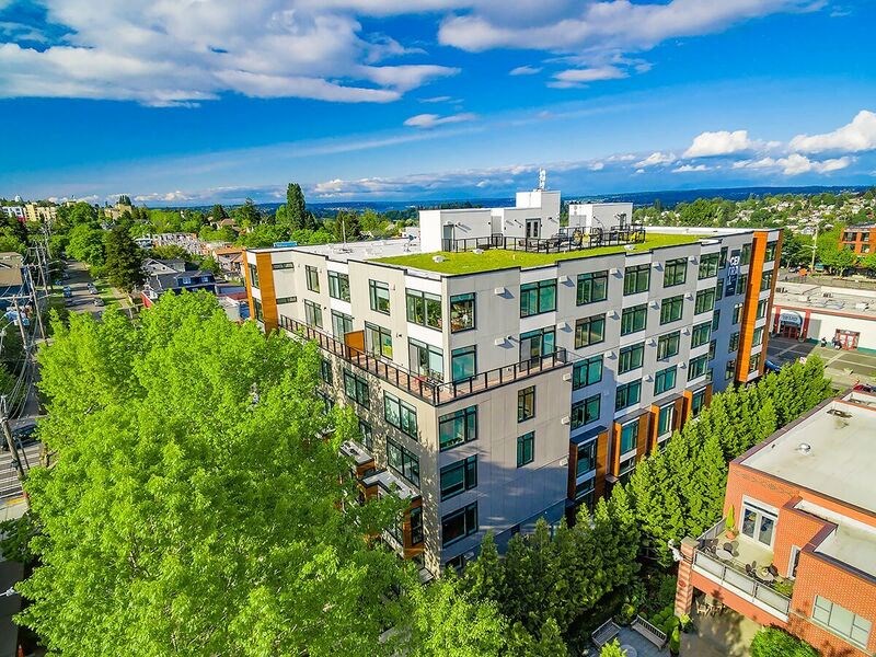 A modern multi-story building with a green roof is surrounded by trees and other buildings.
