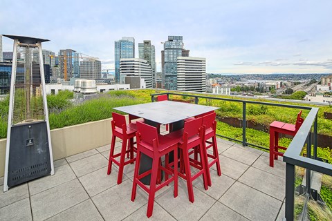A table with red chairs is set up on a balcony with a cityscape in the background.