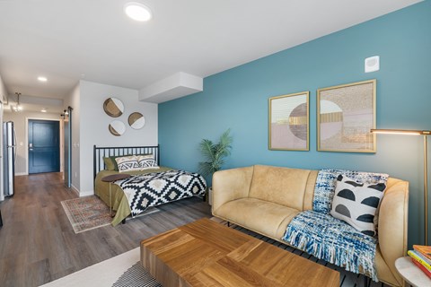 A living room with a tan couch and a bed with a black and white patterned comforter.