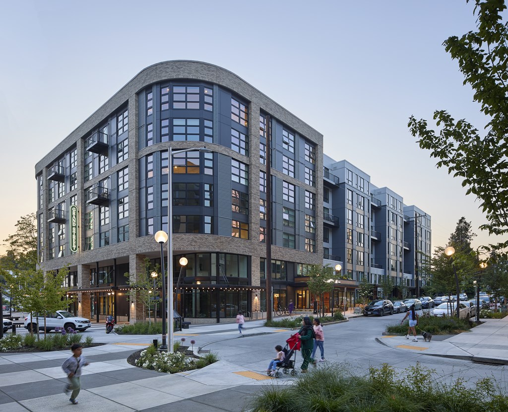 A modern building with a curved facade and large windows is surrounded by a parking lot and people walking on the sidewalk.