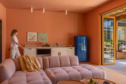 A woman is walking in a living room with a pink couch and a blue refrigerator.