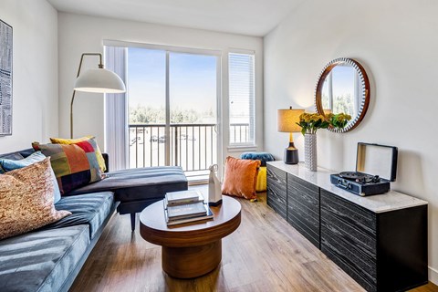 A living room with a grey couch, a round table, a coffee table, a lamp, a mirror, and a balcony with a view of trees.