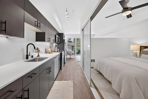 A modern kitchen with a long counter and a bedroom with a bed and a nightstand.