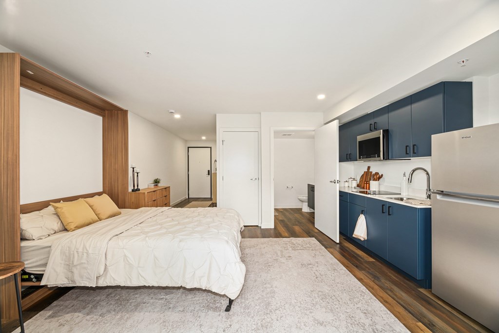 A bedroom with a bed, nightstand, and a kitchen area with a sink and refrigerator.