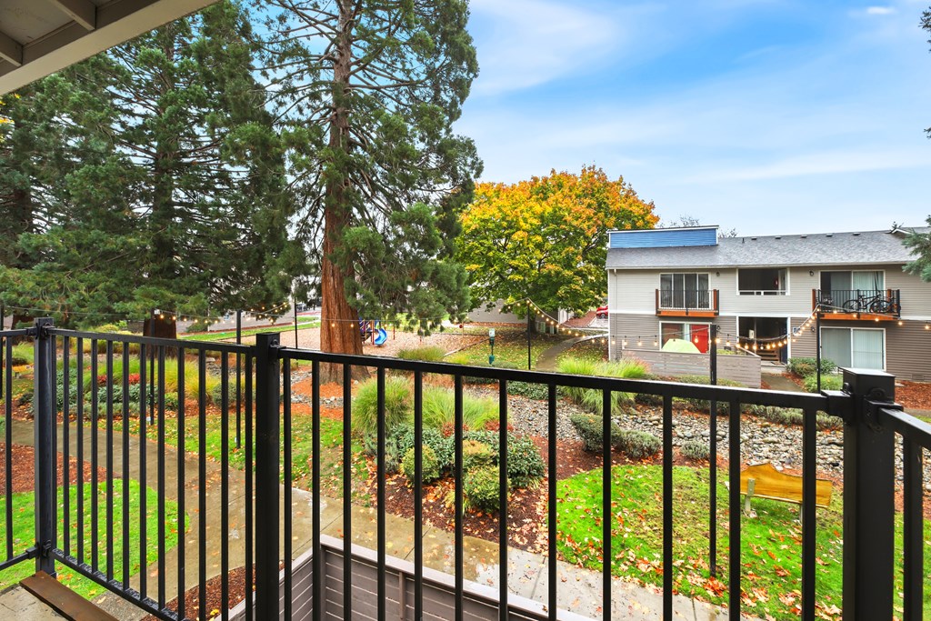 A balcony with a black railing overlooks a garden with a tree and a house in the background.