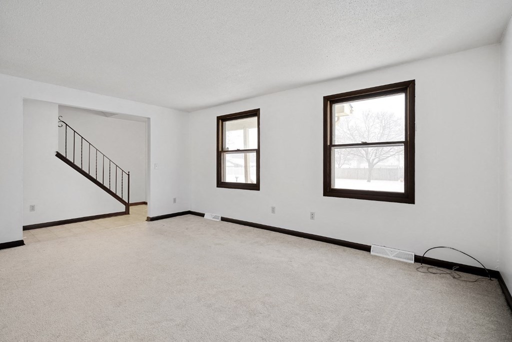 A room with a carpeted floor, a staircase, and two windows.