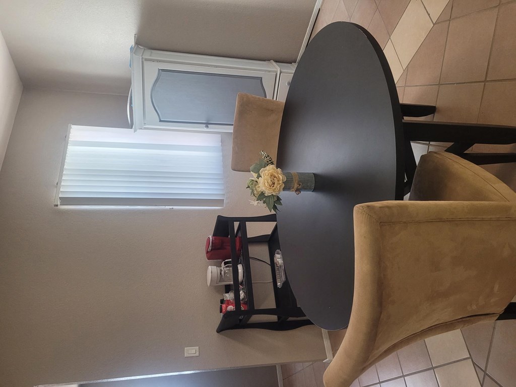 A black chair with a flower on it is in the foreground of a room with a window and some shelves.