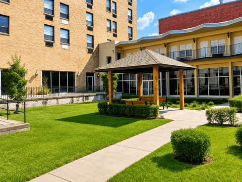A gazebo is in the middle of a grassy area in front of a tan building.