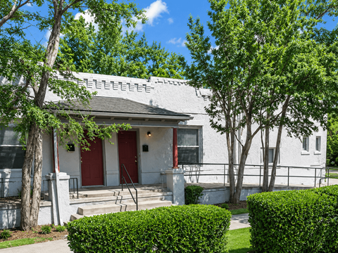 A white house with red doors and windows is surrounded by green bushes and trees.
