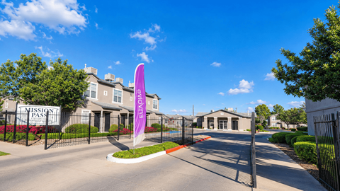 The image shows a gated entrance to a residential area named "Mission Pass" with a purple flag in the foreground.