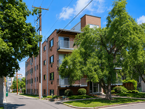 A red brick building with a balcony on the second floor is surrounded by green trees.