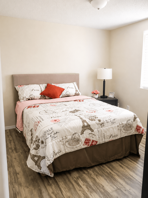A bed with a white comforter and a pink pillowcase is in a room with a lamp and a picture frame on a nightstand.