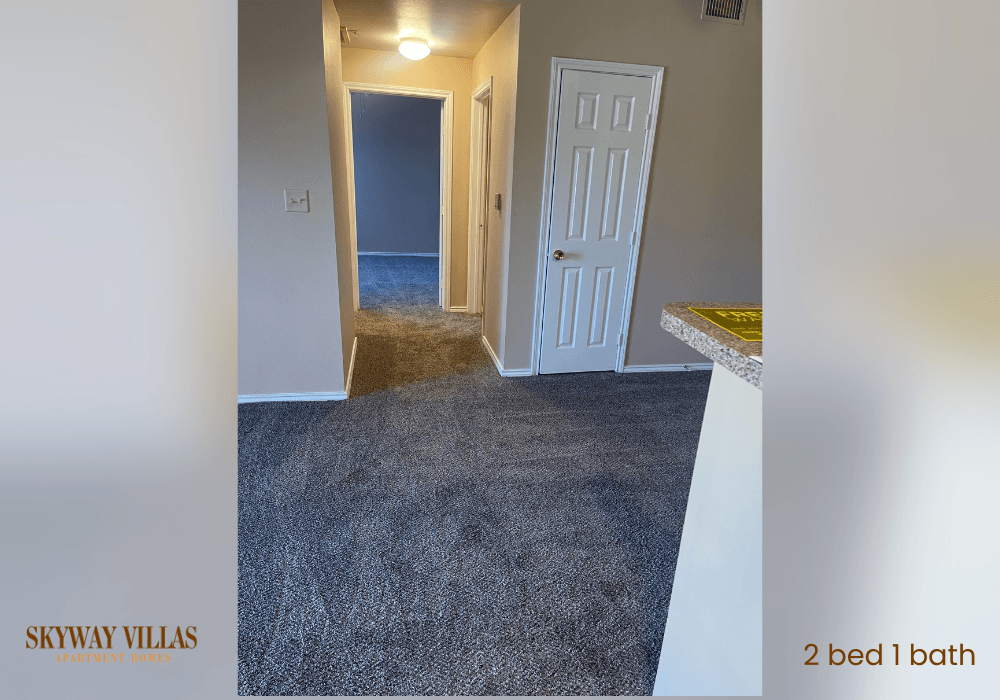 a room with a carpeted floor and a door to a hallway with a kitchen