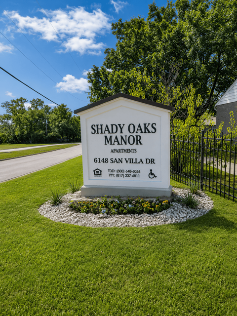 A sign for Shady Oaks Manor Apartments is displayed in front of a fence.