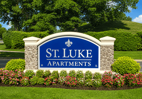 A sign for St. Luke Apartments is surrounded by flowers and greenery.