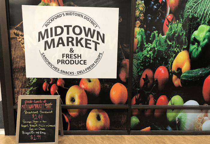 A sign for Midtown Market is displayed in front of a wall of vegetables.