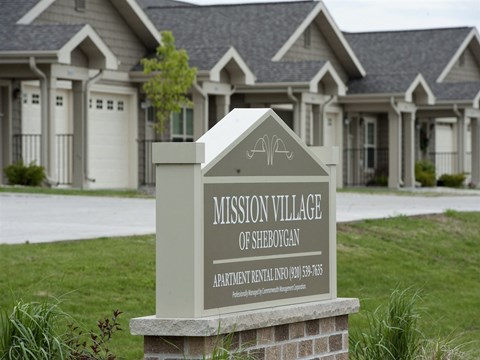 A sign for Mission Village of Sheboygan stands in front of a building.