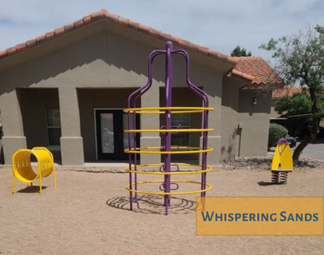 A playground with a purple and yellow structure in front of a building with the words "Whispering Sands" on it.