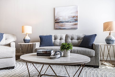 A living room with a grey couch, a grey armchair, a round table with a plant on it, and a painting on the wall.