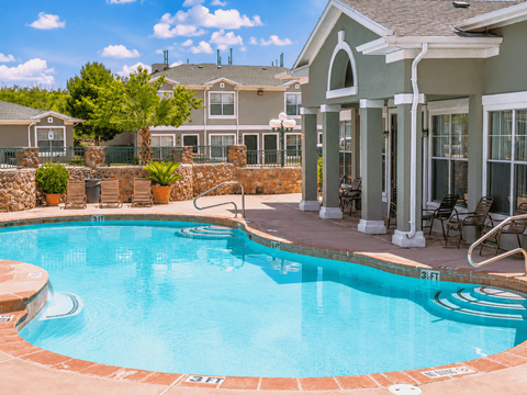 A pool with a stone border and a house in the background.