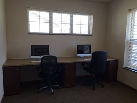 Two computers are on a desk in a room with two windows.