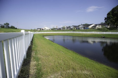 A white fence runs along the edge of a grassy field with a pond in the middle.