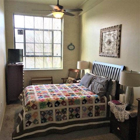 A bedroom with a bed, a ceiling fan, a chair, a dresser, a television, and a window with blinds.