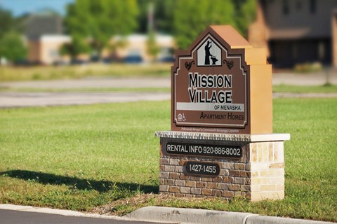 A brown sign that says Mission Village of Menasha in front of a grassy area.