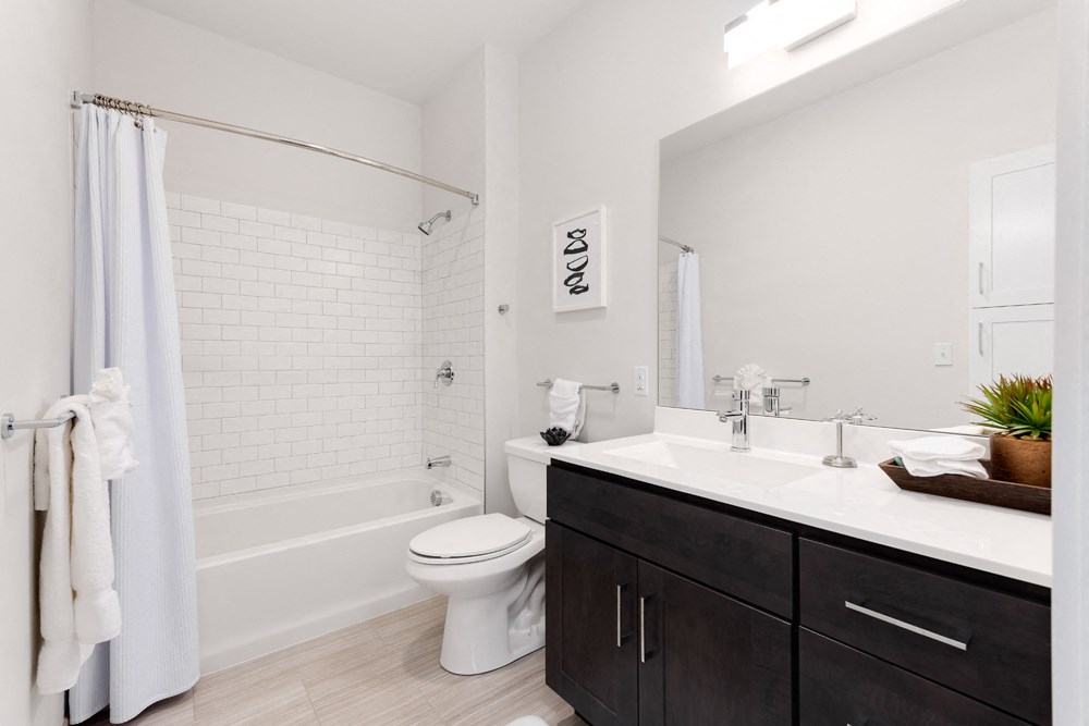 Model apartment bathroom with large mirror, white counter and black cabinet.