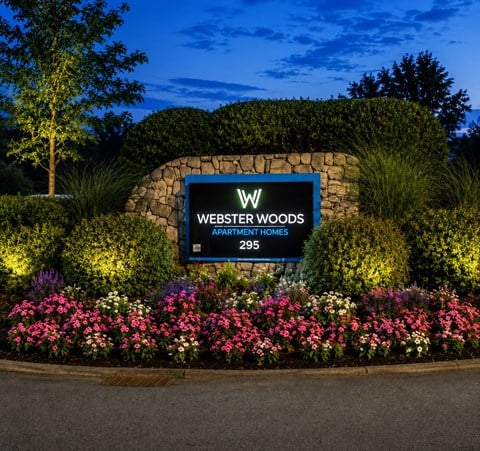 A sign for Webster Woods Apartment Homes is surrounded by flowers and greenery.