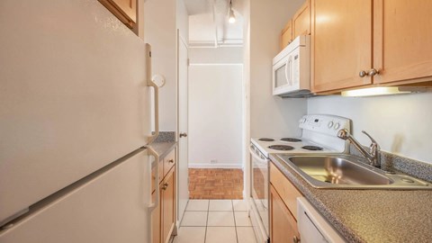 Galley kitchen with white appliances, speckled countertops, light wood cabinetry, and tile flooring.
