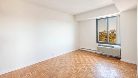 Empty bedroom with parquet flooring, white walls, and large window featuring skyline views at 929 Mass Apartments in Cambridge MA