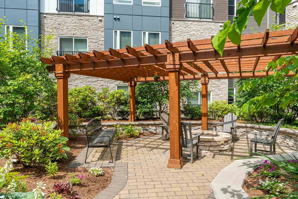 The Residences at Crosspoint central courtyard at 42 Wellman Street in Lowell, MA features paved walkways, raised garden beds, green shrubs, benches, and surrounding multi-story buildings clad in stone and metal siding.