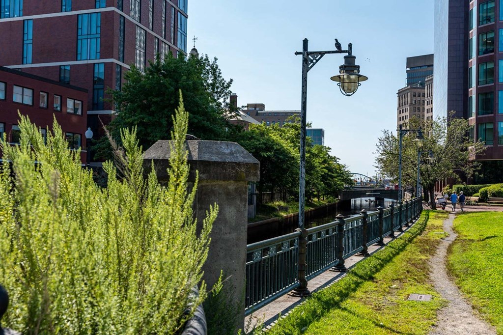 a path next to a river with tall buildings in the background